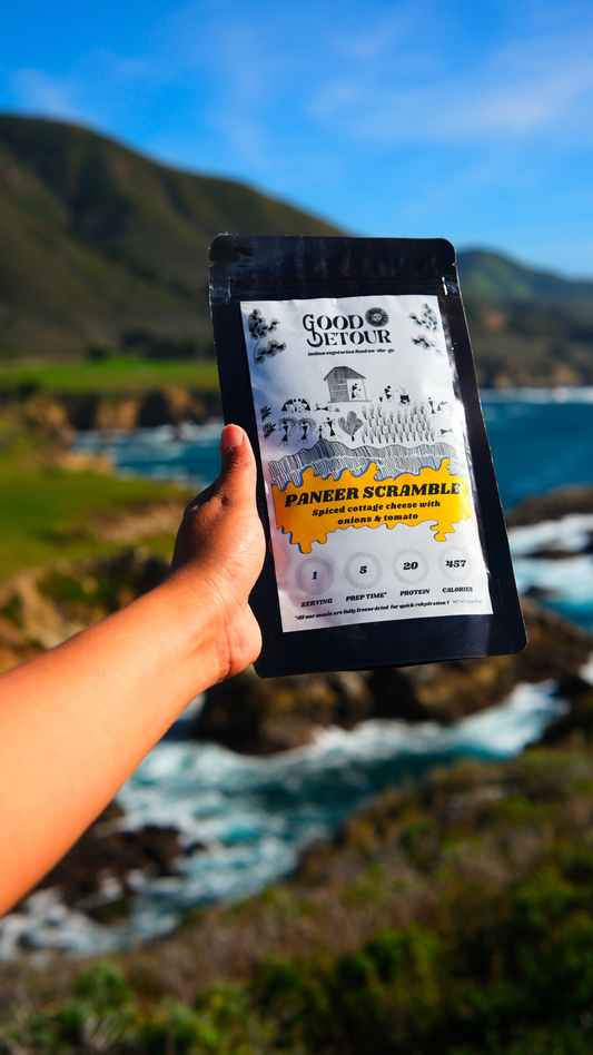 Hand holding a vegetarian Good Detour Paneer Scramble meal pouch with a scenic background of mountains and water.

